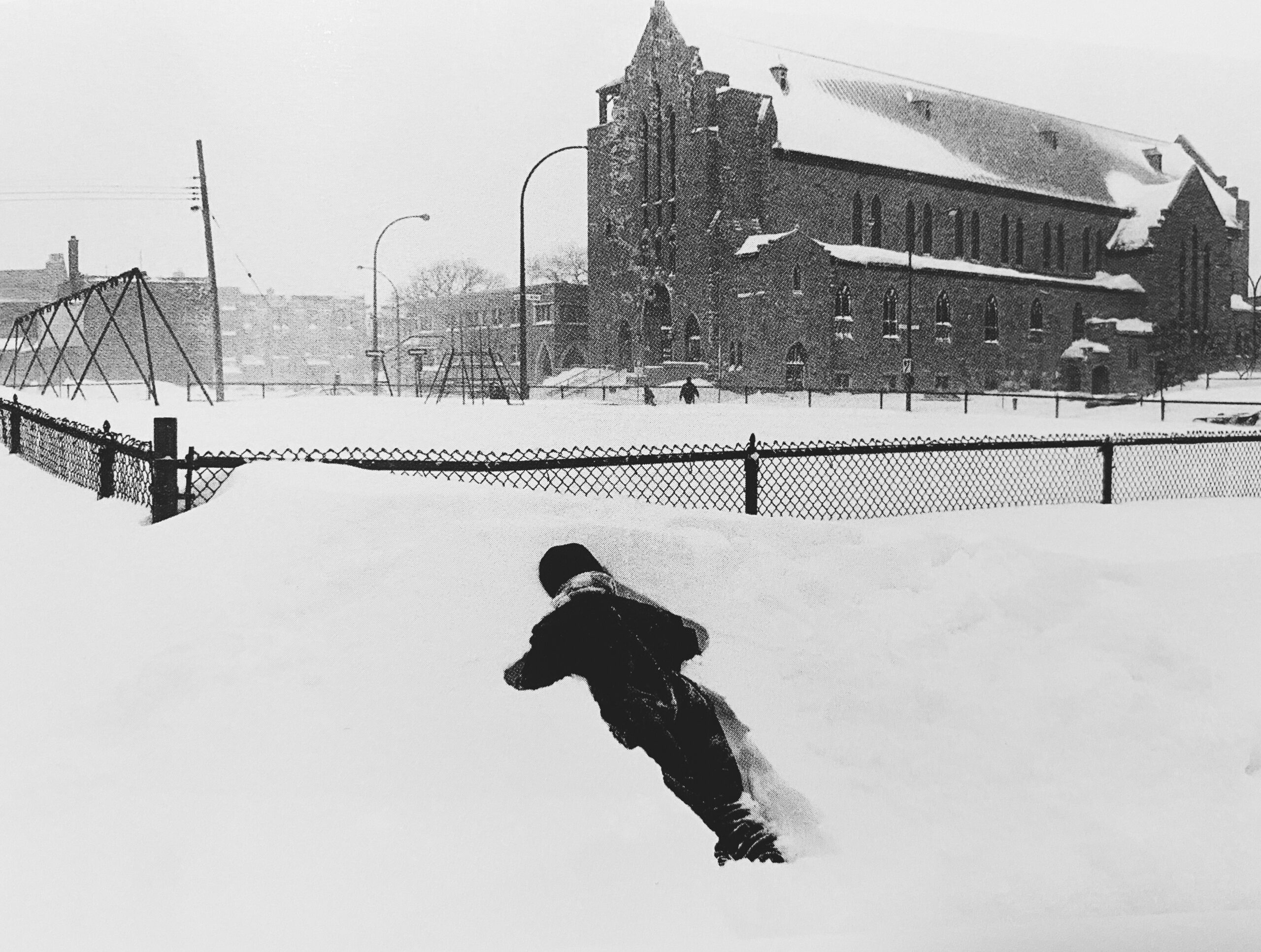 Photographie noir et blanc d'un enfant habillé d'habits d'hiver est étendu dans un bac de neige. Derrière, une clôture enneigée et une église.